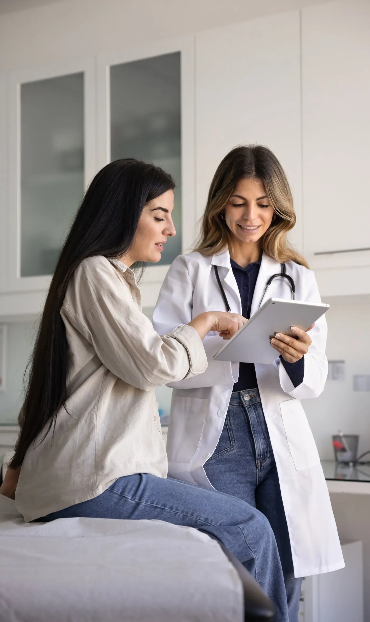 A clinician in a white coat shows a patient something on a tablet in an exam room.