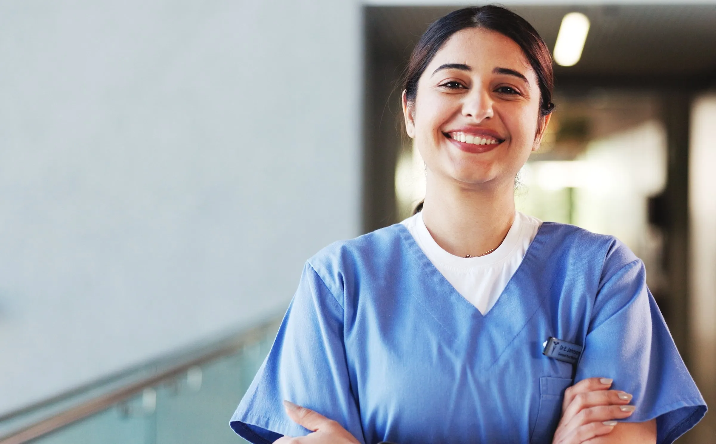 A smiling nurse in blue scrubs stands with arms crossed.